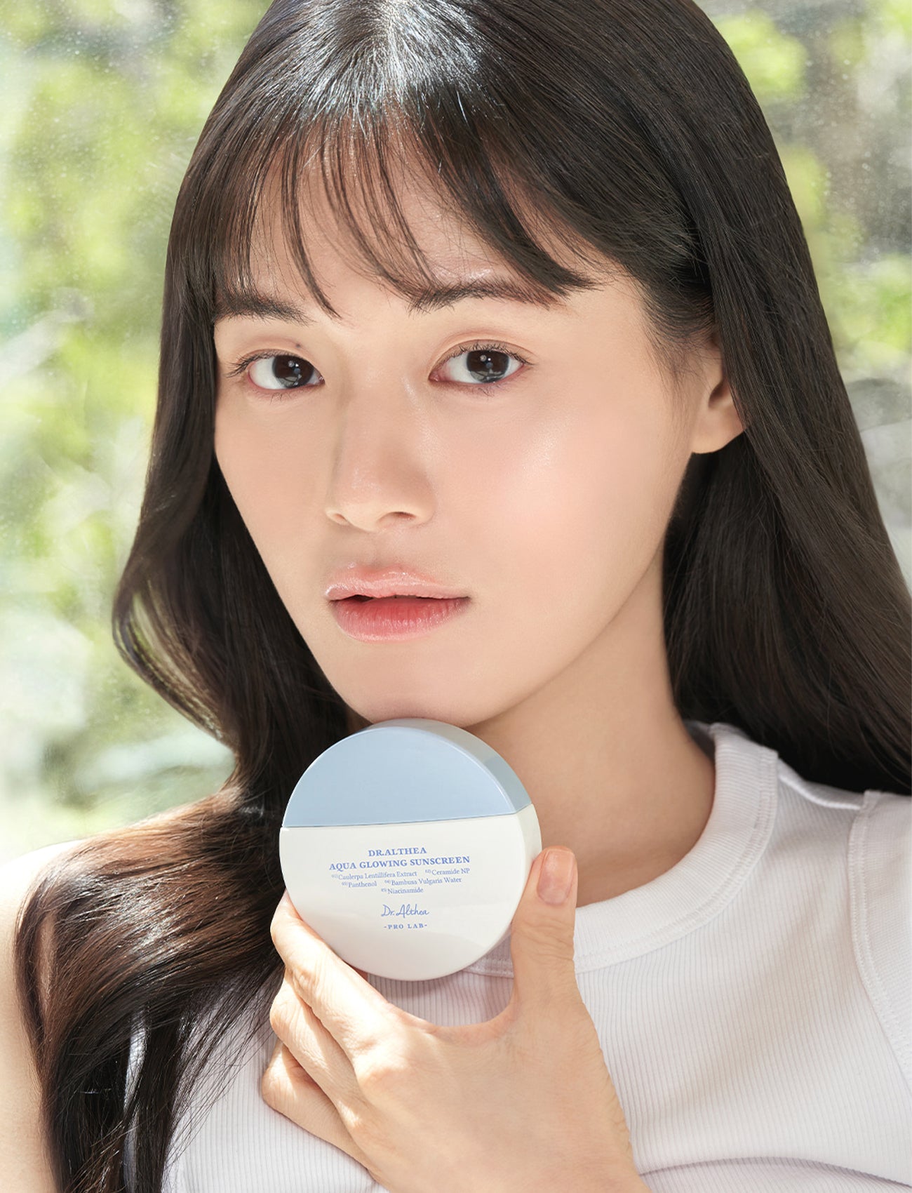 Junge Frau mit glowy Haut hält die Dr. Althea Aqua Glowing Sunscreen vor ihr Gesicht, natürliches Tageslicht im Hintergrund.