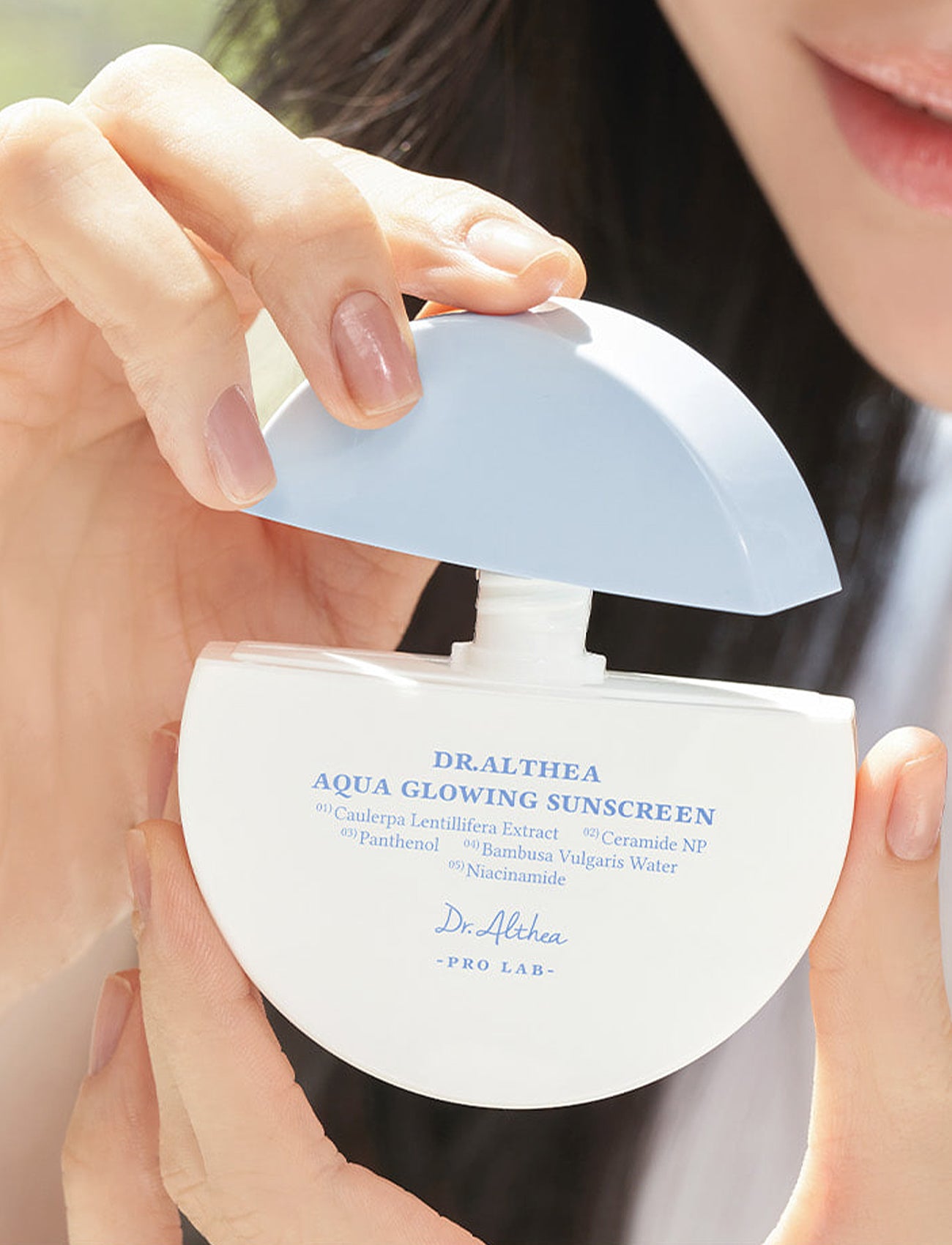 Nahaufnahme einer Hand, die den hellblauen Deckel der Dr. Althea Aqua Glowing Sunscreen abnimmt, Pumpspender sichtbar.