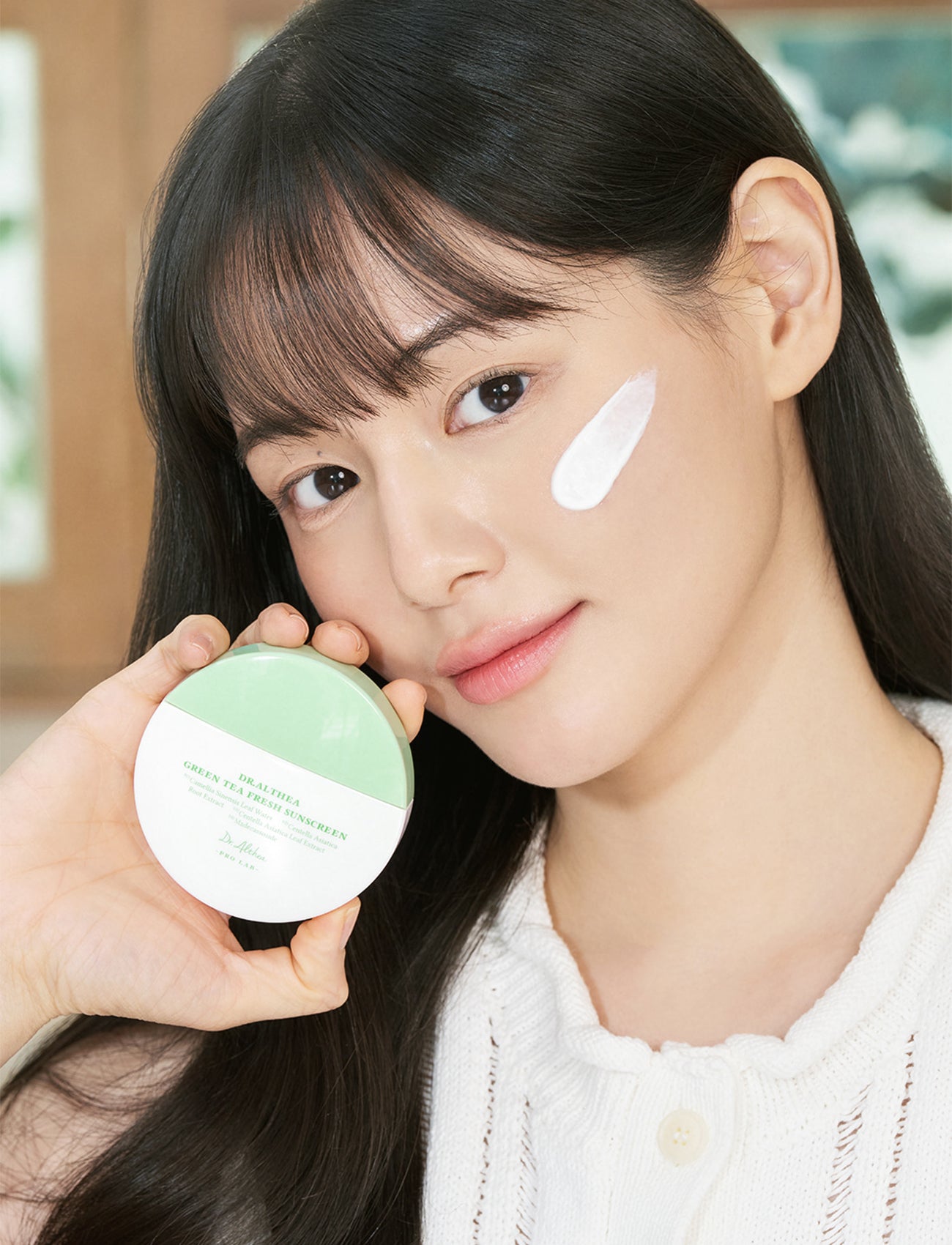 Frau mit einem Streifen Sonnencreme auf der Wange hält die Dr. Althea Green Tea Fresh Sunscreen in die Kamera und lächelt dezent.