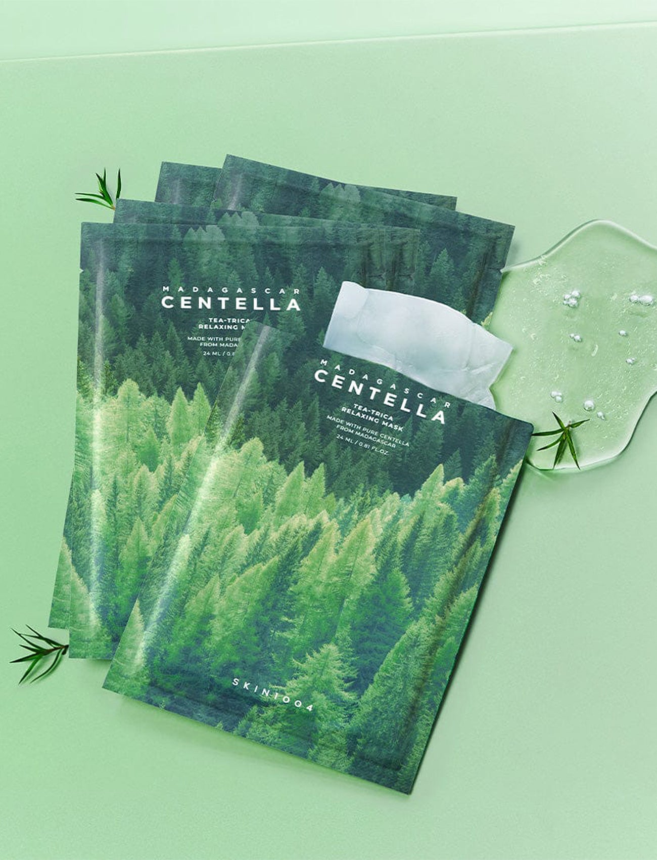 Mehrere SKIN1004 Madagascar Centella Tea-Trica Relaxing Masken nebeneinander auf grünem Untergrund, daneben Geltextur und Teebaumblätter als Hinweis auf die Inhaltsstoffe.